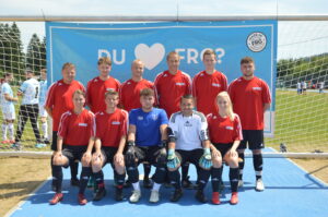 Eine Fußballmannschaft in roten Trikots steht vor einem Tor mit einem Banner im Hintergrund.