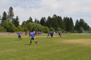 Zwei Mannschaften spielen Fußball auf einem Rasenplatz mit Bäumen im Hintergrund.