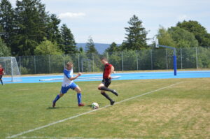 Zwei Fußballspieler, einer in blau-weißer und der andere in rot-schwarzer Uniform, kämpfen um den Ball auf einem Rasenplatz.