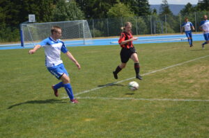 Ein Fußballspieler in blauem Trikot läuft dem Ball hinterher, während ein Spieler in rotem Trikot ihn verfolgt.