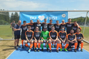 Eine Fußballmannschaft in orange-schwarzen Trikots steht vor einem Tor mit einem Banner im Hintergrund.
