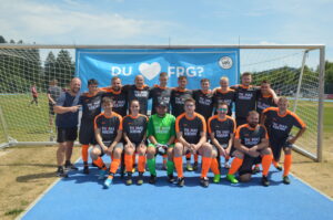 Eine Fußballmannschaft in orange-schwarzen Trikots steht vor einem Tor mit einem Banner im Hintergrund.