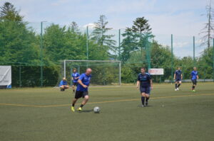 Zwei Spieler in blauen Trikots spielen Fußball auf einem Sportplatz mit Kunstrasen.