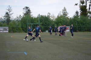 Zwei Fußballspieler führen einen Pass auf einem Sportplatz aus, während andere Spieler zuschauen.
