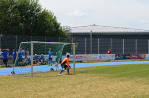 Ein Torwart in orangefarbener Kleidung steht vor einem Fußballtor auf einem Sportplatz.
