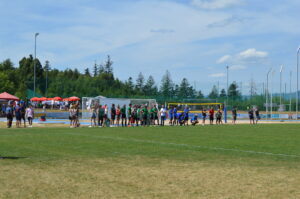 Gruppen von Spielern und Zuschauern auf einem Fußballplatz während eines Spiels unter blauem Himmel.