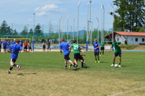 Fußballspiel mit Spielern in blauen und grünen Trikots auf einem Rasenfeld.