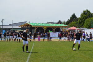 Zwei Fußballspieler in schwarzer Ausrüstung stehen auf dem Spielfeld, während Zuschauer im Hintergrund stehen.