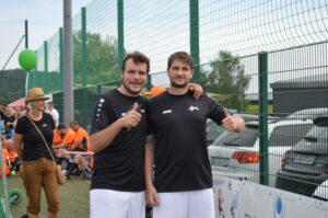 Zwei Männer in schwarzen Fußballtrikots stehen nebeneinander und lächeln in die Kamera.