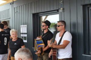 Zwei Musiker spielen Akkordeon und Gitarre in einem Innenraum mit Zuschauern im Hintergrund.