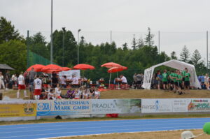 Gruppe von Zuschauern und Sportlern bei einer Veranstaltung im Freien mit Zelten und Sonnenschirmen.