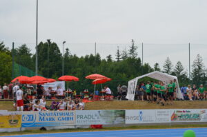 Menschen auf einer Sportveranstaltung mit roten Sonnenschirmen und Zelten im Hintergrund.