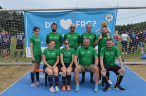 Eine Gruppe von neun Personen in grünen Trikots steht vor einem Fußballtor mit einem Banner im Hintergrund.