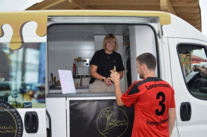 Eine Verkäuferin im Food Truck spricht mit einem Kunden, der ein rotes Trikot trägt.