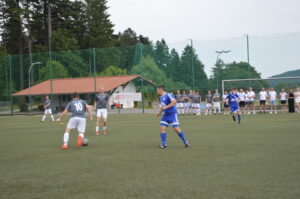 Zwei Fußballspieler, einer in grauem und einer in blauem Trikot, spielen auf einem Rasenplatz.