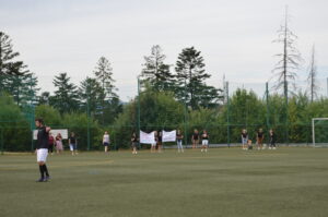Fußballspieler auf dem Feld mit Zuschauern, die Transparente halten, im Hintergrund Bäume.