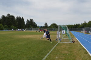 Ein Fußballspieler läuft am Tor auf einem Sportplatz mit anderen Spielern im Hintergrund.
