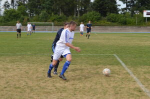 Zwei Fußballspieler, einer in einem weißen Trikot und der andere in einem blauen Trikot, kämpfen um den Ball auf dem Spielfeld.