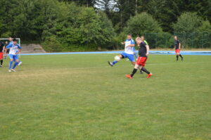 Zwei Spieler in blauen und roten Trikots, die auf einem Rasenplatz Fußball spielen.