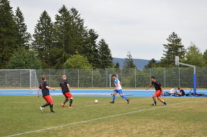 Drei Spieler in schwarzen und roten Trikots spielen Fußball auf einem Sportplatz mit einem Tor im Hintergrund.