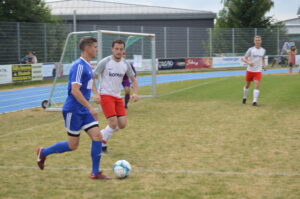 Zwei Fußballspieler, einer in blauem und einer in rotem Trikot, spielen auf einem Rasenplatz.