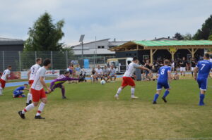 Fußballspiel mit Spielern in roten und blauen Trikots auf einem Sportplatz.