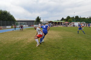 Zwei Fußballspieler, einer in einem roten und der andere in einem blauen Trikot, kämpfen um den Ball auf einem Spielfeld.