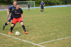 Ein Spieler in schwarzem Trikot mit roten Shorts dribbelt den Ball, während ein blauer Spieler ihn verfolgt.