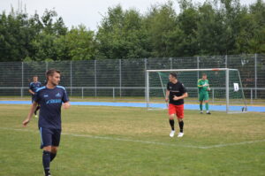 Zwei Fußballspieler auf dem Feld, einer in blauem Trikot und der andere in rotem Trikot, mit einem Tor im Hintergrund.