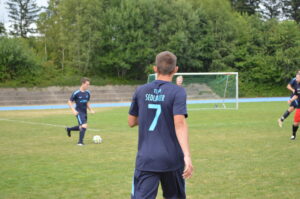 Zwei Fußballspieler in blauen Trikots auf einem Spielfeld mit Tor im Hintergrund.