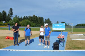 Vier Personen stehen auf einem Sportplatz neben einer Tisch mit Trophäen und einem Banner im Hintergrund.