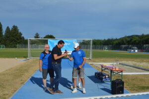 Drei Männer stehen auf einem Sportplatz während einer Veranstaltung zur Ehrung von Teilnehmern.