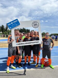 Eine Gruppe von Fußballspielern in grauen Trikots mit orangefarbenen Akzenten posiert vor einem großen Rahmen.