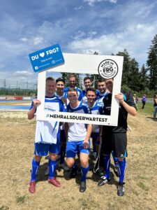 Eine Gruppe von sieben Fußballspielern posiert mit einem großen Rahmen auf einem Sportplatz.