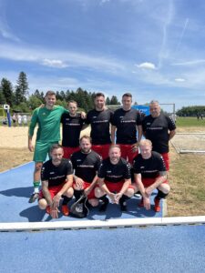Eine Fußballmannschaft in schwarzen und roten Trikots steht auf dem Spielfeld mit einem Tor im Hintergrund.