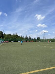Fußballmannschaften spielen auf einem Kunstrasenplatz unter blauem Himmel mit Wolken.