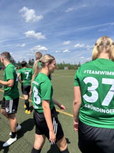 Spielerinnen einer Fußballmannschaft in grünen Trikots mit der Nummer 37 und dem Hashtag #TEAMWIMMER.
