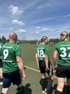 Drei Fußballspieler in grünen Trikots mit den Nummern 9, 18 und 37 auf einem Sportplatz.