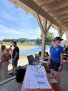 Frau in blauer Sportbekleidung steht an einem Tisch mit Unterlagen und Wasserflasche bei einer Sportveranstaltung.