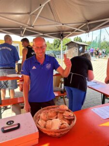 Ein Mann in blauer Sportkleidung zeigt einen Daumen hoch neben einem Korb mit verschiedenen Brötchen.