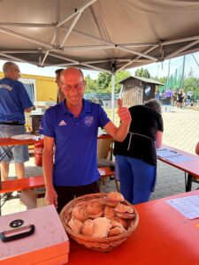 Ein Mann in blauer Sportkleidung zeigt einen Daumen hoch und hält einen Korb mit Brötchen an einem Verkaufsstand.