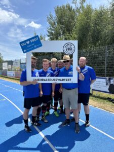 Sechs Fußballspieler in blauen Trikots posieren mit einem Rahmen auf einem Sportplatz.