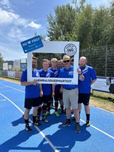 Eine Gruppe von sechs Personen in blauen T-Shirts posiert mit einem großen Rahmen auf einem Sportplatz.
