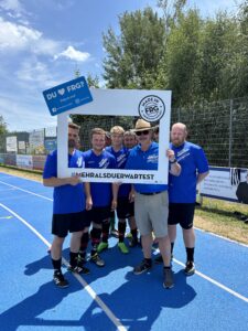 Sechs Fußballspieler in blauen Trikots posieren mit einem Rahmen auf einem Sportplatz.