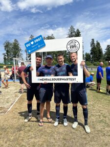 Vier Personen posieren mit einem großen Rahmen auf einem Sportplatz während eines Events.