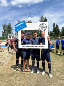 Vier Männer in Sportkleidung halten einen großen Rahmen mit dem Text '#MEHRALSDUERWARTEST'.