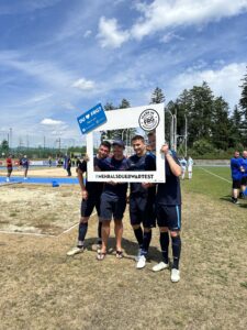 Drei Fußballspieler posieren mit einem großen Rahmen auf einem Sportplatz.