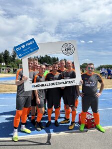 Eine Fußballmannschaft posiert mit einem Rahmen und einem Banner auf einem Sportplatz.