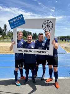 Vier Fußballspieler posieren mit einem großen Rahmen und einem Schild auf einem Sportplatz.