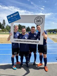 Vier Fußballspieler posieren mit einem großen Rahmen, der den Text 'DU ♥ FRG?' enthält.
